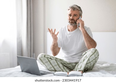 Irritated angry handsome grey-haired middle aged man wearing pajamas sitting on bed at home, using laptop, have phone conversation and gesturing, have argument, copy space - Powered by Shutterstock