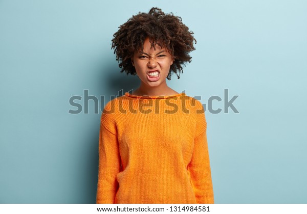 いらいらした黒い若い可愛い女性が顔をしかめ 怒りで歯を食いしばり 否定的な反応を示し 黒い肌をし オレンジのジャンパーを着け 青い背景にモデルを着け ひどい扱いに悩まされる の写真素材 今すぐ編集