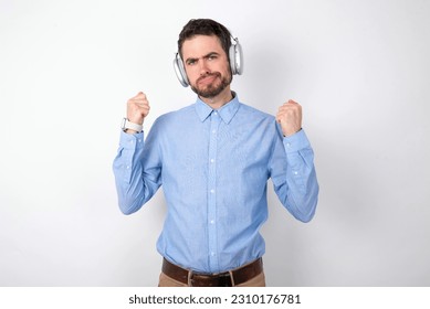 Irritated Businessman wearing blue t-shirt with headphones over white background blows cheeks with anger and raises clenched fists expresses rage and aggressive emotions. Furious model - Shutterstock ID 2310176781