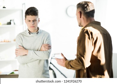 Irritated Father Gesturing And Looking At Teen Son With Crossed Arms At Home