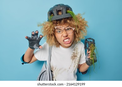 Irritated female leads active lifestyle rides bicycle gestures angrily and clenches teeth wears safety helmet and gloves white dirty t shirt annoyed because of bike breakdown isolated over blue wall