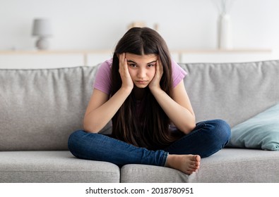Irritated Indian teenage girl sitting on couch, feeling bored, suffering from lack of communication, being trapped at home during coronavirus lockdown, having teen depression, full length