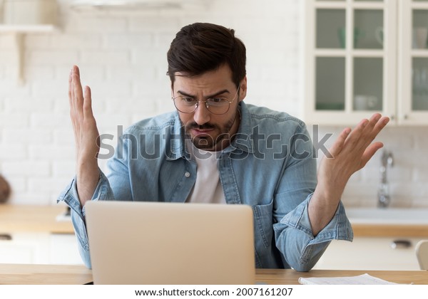 Irritated man in glasses sit at table staring\
at laptop screen spread arms feels angry and confused having\
problem with broken computer, reading bad news in email, lost\
information, app\
malfunction