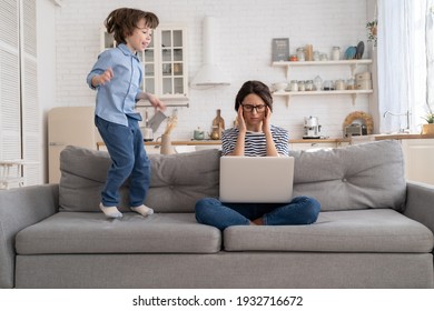 Irritated Mother Sitting On Couch Working On Laptop At Home, Distracted From Noise And Touching Her Temple, Hyperactive Little Kid Jumping On Sofa And Drawing Attention. Tired Mom Can't Concentrate.