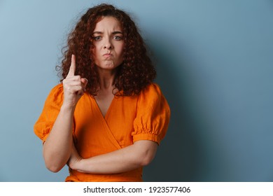 Irritated redhead curly girl wagging her finger at camera isolated over blue background