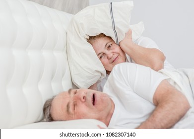 Irritated senior woman blocking ears with pillow while her husband snoring on bed - Powered by Shutterstock