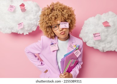 Irritated sulking female office worker looks directly at camera dressed in formal clothes uses sticky notes to remember important tasks to do poses with folders isolated over pink background - Shutterstock ID 2255389311