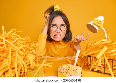 Irritated Woman Bites Lips And Scratches Head Poses At Desktop With Cut Paper Around Fed Up Of Long Hours Working Eats Cereals For Breakfast Isolated Over Yellow Background. Annoyed Office Worker