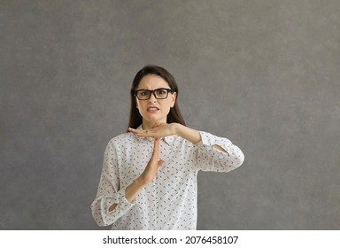 Irritated woman in glasses against grey studio background looking at you, doing time out sign hand gesture, asking to end unpleasant conversation or warning that you need to stop abusive relationship