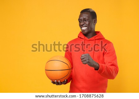 Irritated young african american man guy basketball player in red streetwear hoodie posing isolated on yellow background. Sport leisure lifestyle concept. Playing basketball hold ball clenching fist