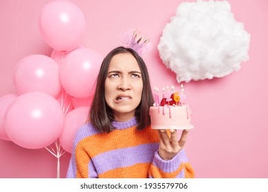 Irritated Young Asian Woman With Dark Hair Bites Lips Being Displeased About Getting Older Wears Princess Crown On Head Holds Festive Cake Poses Near Decorated Background Fed Up Of Holidays.