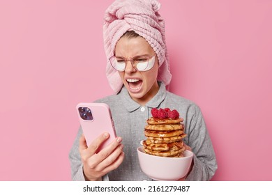 Irritated young woman yells angrily focused at smartphone scrolls newsfeed wears towel on head and slumber suit prepared appetizing pancakes with rasberry for family breakfast feels outraged