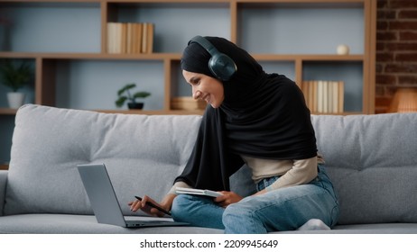 Islamic Arabian Student Girl Muslim Woman Wearing Black Hijab Busy Lady In Headphones With Laptop Watching Webinar Lesson Online Class Training Write Notes In Notebook Do Homework E-learning At Home