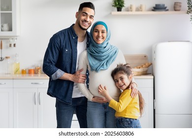 Islamic Expecting Family. Happy Pregant Muslim Woman In Hijab Posing With Husband And Little Daughter, Arab Spouses And Cute Female Child Standing In Kitchen Interior And Smiling At Camera