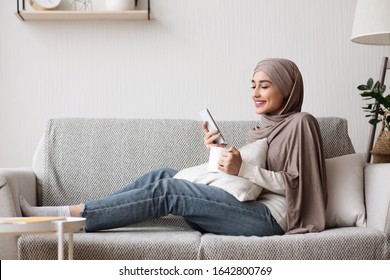 Islamic Housewife Leisure Concept. Young Muslim Woman In Hijab Relaxing At Home With Smartphone And Coffee, Side View - Powered by Shutterstock