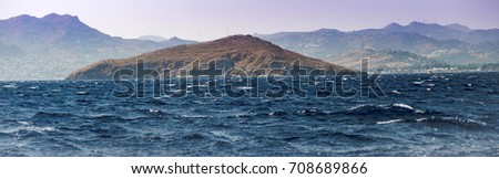 An island at Aegean Sea on a choppy waters with foggy land in the background.