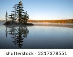 An island reflects on a calm lake with an autumn colored, tree lined shoreline and the moon in the background. Islet Lake, Algonquin Provincial Park, Ontario, Canada.