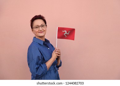 Isle Of Man Flag. Business Woman Holding Isle Of Man Flag. Nice Portrait Of Middle Aged Lady 40 45 Years Old With A National Flag Over Pink Wall On The City Street Outdoors.