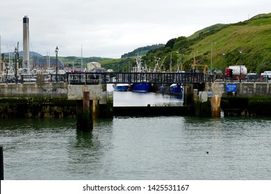 Isle Of Man UK June 2019. Peel Town. Looking Towards The Pedestrian Swing Bridge And Metal Dam That Guards The Inner Marina. Tower Of Waste Disposal Factory In Background. Boats Moored In Marina. 