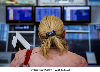 Isolated Back View Of A Blonde Woman With A Pony Tail In A Pink Coat Holding An  Airplane Ticket, A Passport, Searching For A Flight Time On A Blurred Blue Departure Board In A Terminal Of An Airport.