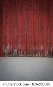 Isolated Backdrop Of A Red Barn Wood Wall And Gray Cement Flooring. Bottoms Of Vertical Wooden Planks Are Distressed, Worn, Fraying And Dark With Age. Slightly Darkened Vignette.