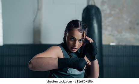 Isolated Badass Confident Female Kick Boxer Looking Directly at the Camera As She Strikes a Fight Move Pose. Training Hard in an Old Boxing Studio Gym With Punching Bags. Athletic Martial Arts Woman.