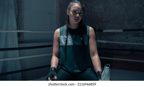 Isolated Badass Confident Looking Female Kick Boxer Standing In The Middle Of A Boxing Ring, Holding A Water Bottle And Looking Directly At The Camera. Ready To Start Training For Championship.