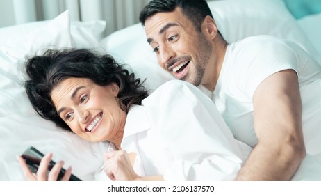 Isolated Beautiful Couple Laying In Bed Celebrating Valentines Day By Staying In A Fancy Hotel Suite. Ethnic Woman Holding A Smartphone Device Taking Selfies Together, Laughing At How They Turned Out.