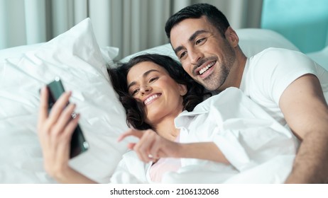 Isolated Beautiful Ethnic Couple Laying In Bed In A Fancy Hotel Room, Dressed In All White, Woman Holding A Smartphone Device Taking Selfies Together, Smiling And Having A Good Time.