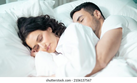 Isolated Beautiful Ethnic Couple Laying In Bed Cuddling Together Wearing All White, Sleeping In Celebrating The Valentines Day Together In A Fancy Hotel Suite. Valentines Day Concept.