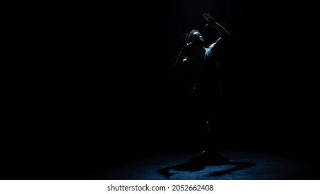 Isolated Beautiful Ethnic Male Ballet Dancer Showing A Dance Move With Hands In The Sky, The Light Reflecting Off Of His Face. Background Is A Dark And Hazy Theatre With A Stage.