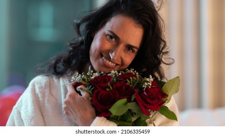 Isolated Beautiful Ethnic Woman Looking Directly At The Camera Holding A Bouquet Of Roses After Getting Proposed To, Sitting On The Bed Of A Fancy Hotel Suite Room Celebrating Valentines Day.