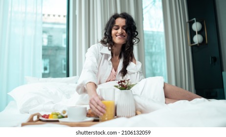 Isolated Beautiful Ethnic Woman Sitting On The Bed Of A Fancy Hotel Suite Grabbing An Orange Juice Glass Off Of A Breakfast Tray, Big Windows In The Background. Celebrating Valentines Day.