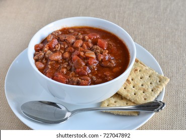 Isolated Bowl Of Chili Soup With Spoon And Whole Wheat Saltine Crackers.  Beans, Beef, Tomatoes, And Spices As Main Ingredients.