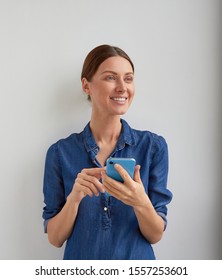 Isolated Brown Tucked Back Hair Woman With Happy Face In Blue Jeans Shirt Dress Uses Phone Holding By Two Hands With Wide Beautiful Smile With White Teeth Looks To Side To Hear Someone