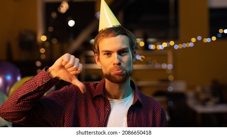 Isolated Caucasian Man Standing In An Office, Wearing A Party Hat And Celebrating Co-Workers Birthday Party. Looking Directly At The Camera And Showing A Thumbs Down With A Sad Face.