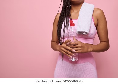 Isolated close-up portrait on pink background of an unrecognizable African woman in pink tracksuit and towel holding a bottle with water, refreshing herself after workout. Copy ad space