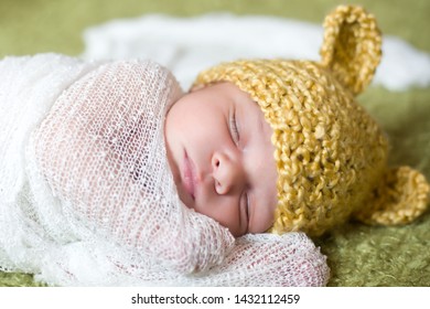 Isolated Closeup Of A Week-old Baby Boy, Light Skinned (mixed African And Caucasian Ethnicities) Posed Lying On His Side Wrapped In A White Knit Blanket And Wearing A Crocheted Yellow Bear Hat.