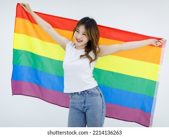 Isolated Cutout Full Body Studio Shot Of Millennial Asian Young Happy Female Lesbian LGBTQ Model In Casual Outfit Standing Smiling Holding Rainbow Pride Proud Unity Equality Flag On White Background.