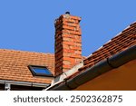 Isolated damaged clay brick chimney with weathered and spalling surface. clay tile roofing. metal flashing. strong shadow. blue sky background, summer scene. 