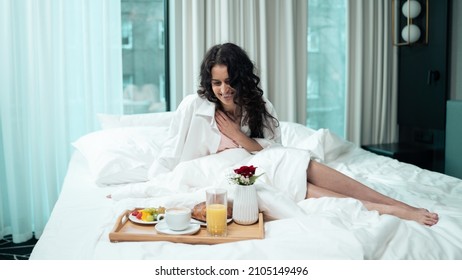 Isolated Ethnic Female Sitting On The Bed Of A Fancy Hotel Suite, Wearing A White Shirt And Pink Pyjamas, Waking Up In The Morning Looking Fresh And Well Rested, Cute Breakfast Tray In Front Of Her.