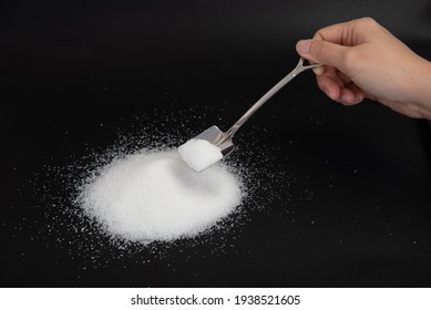 Isolated Hand Holding A Shovel Shaped Silver Spoon Scooping A Pile Of Pure White Sugar On Black Background