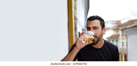 Isolated Hispanic Latin Arab Indian Bearded Handsome Guy Drinking Beer With A A Blurred Colonial City Background, And Plenty Of Horizontal Copy Space. Ads, Banner, Blank Space And Advertising Concept