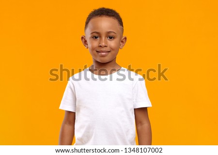 Isolated image of cute adorable dark skinned schoolboy wearing white t-shirt posing in blank yellow studio with confident cheerful smile because he received high marks for maths test at school