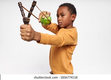 Isolated image of serious concentrated little black boy holding Y-shaped stick with elastic, shooting green apple, having focused facial expression. Accurate African child playing with slingshot