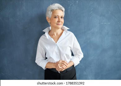 Isolated image of stylish experienced middle aged female executive with short gray hair standing in confident posture, wearing elegant black trousers and white formal shirt, smiling at camera