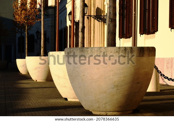 isolated large white concrete tree planter pots in\
diminishing perspective. small trees in brown autumn leaf colors.\
stucco facade and sctreetscape. antique or vintage stile street\
light or sconce 