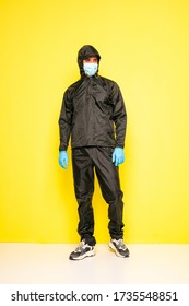 Isolated Man On Yellow Background In Full Body Black Costume, With Medical Mask And Steril Gloves Posing To Camera