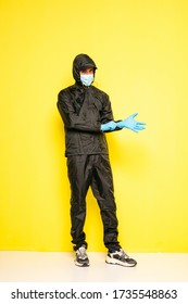 Isolated Man On Yellow Background In Full Body Black Costume, With Medical Mask And Steril Gloves Posing To Camera