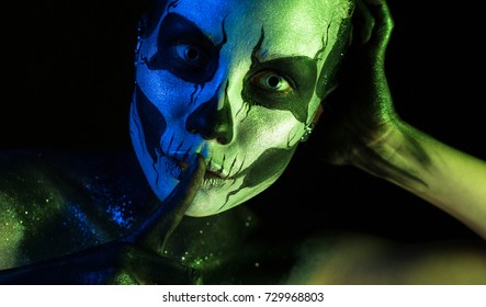 Isolated on black, closeup picture, toned green and blue, beautiful 
creepy young blonde caucasian woman with scull body art, grey eyes, hand on head, points to be silent, look at camera